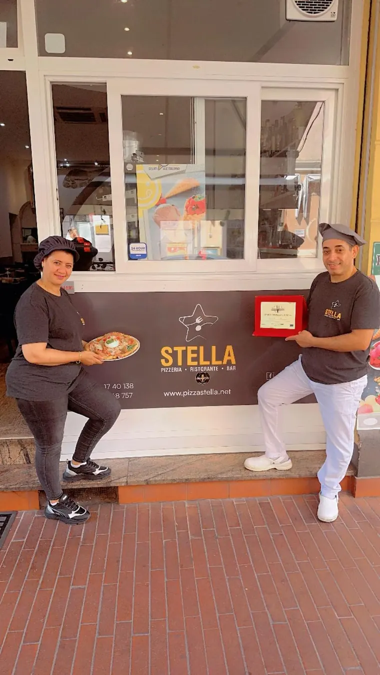 Due persone dello staff di Stella davanti al locale, in maglietta Stella: una tiene una pizza, l'altra la targa del premio